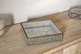 The nkuku Malita Jewellery Box in antique brass rests on a grooved wooden surface, holding a gold bracelet and necklace. Nearby are a candle in a frosted holder and another mercury glass jewellery box, set against a light wall.