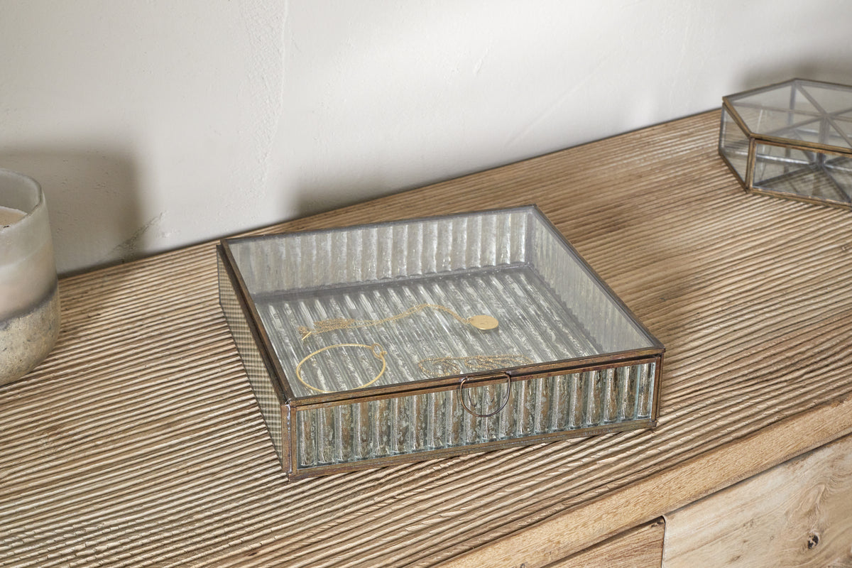 The nkuku Malita Jewellery Box in antique brass rests on a grooved wooden surface, holding a gold bracelet and necklace. Nearby are a candle in a frosted holder and another mercury glass jewellery box, set against a light wall.