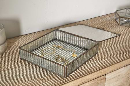 The Malita Jewellery Box - Antique Brass by nkuku sits open on a textured wooden dresser, displaying a gold bracelet, necklace, and earrings. Its ribbed glass lid is partially lifted, with a candle placed nearby.