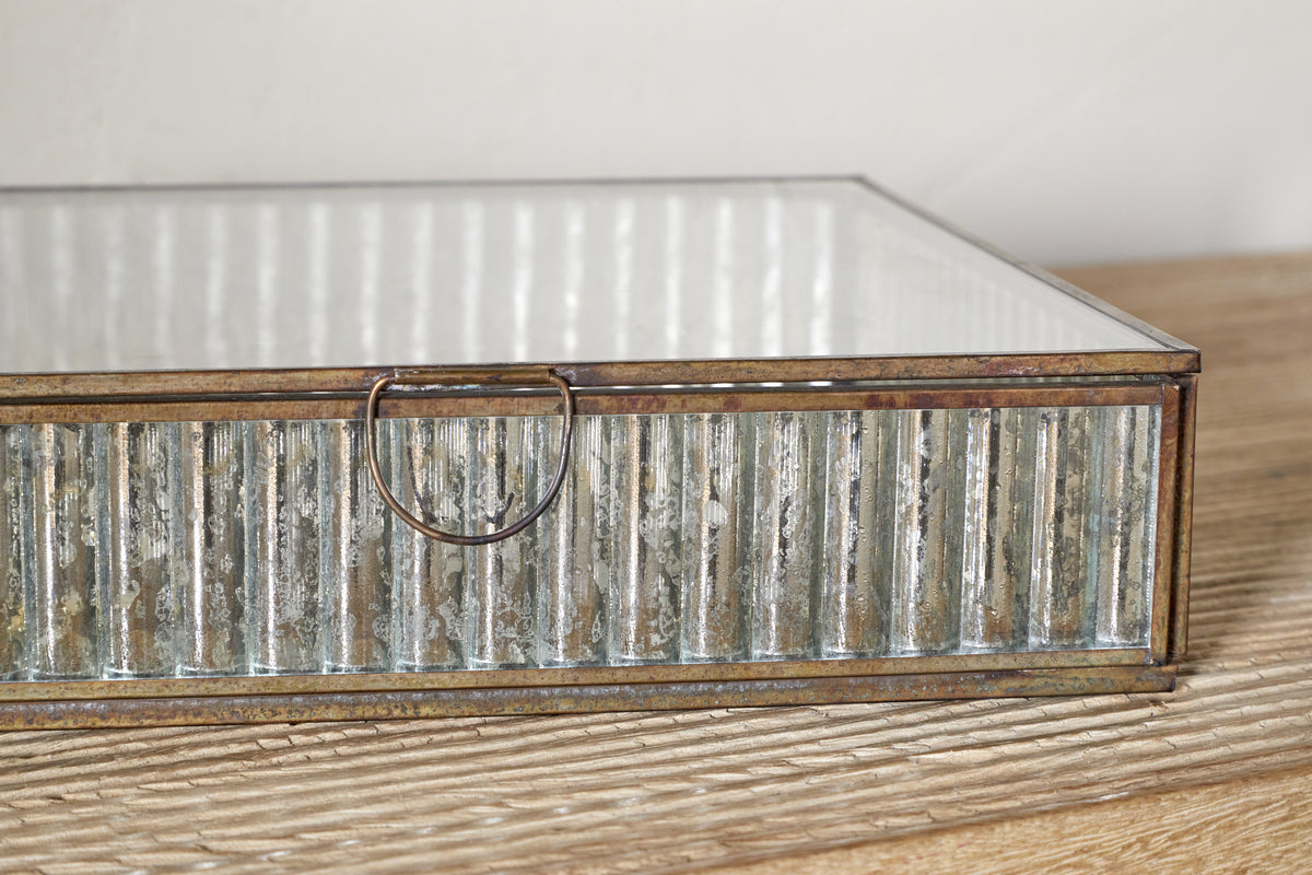 A close-up of the Malita Jewellery Box by nkuku, styled in antique brass with a mirrored base and mercury glass-inspired sides. It features a small circular handle and is set on light wood against a neutral background.
