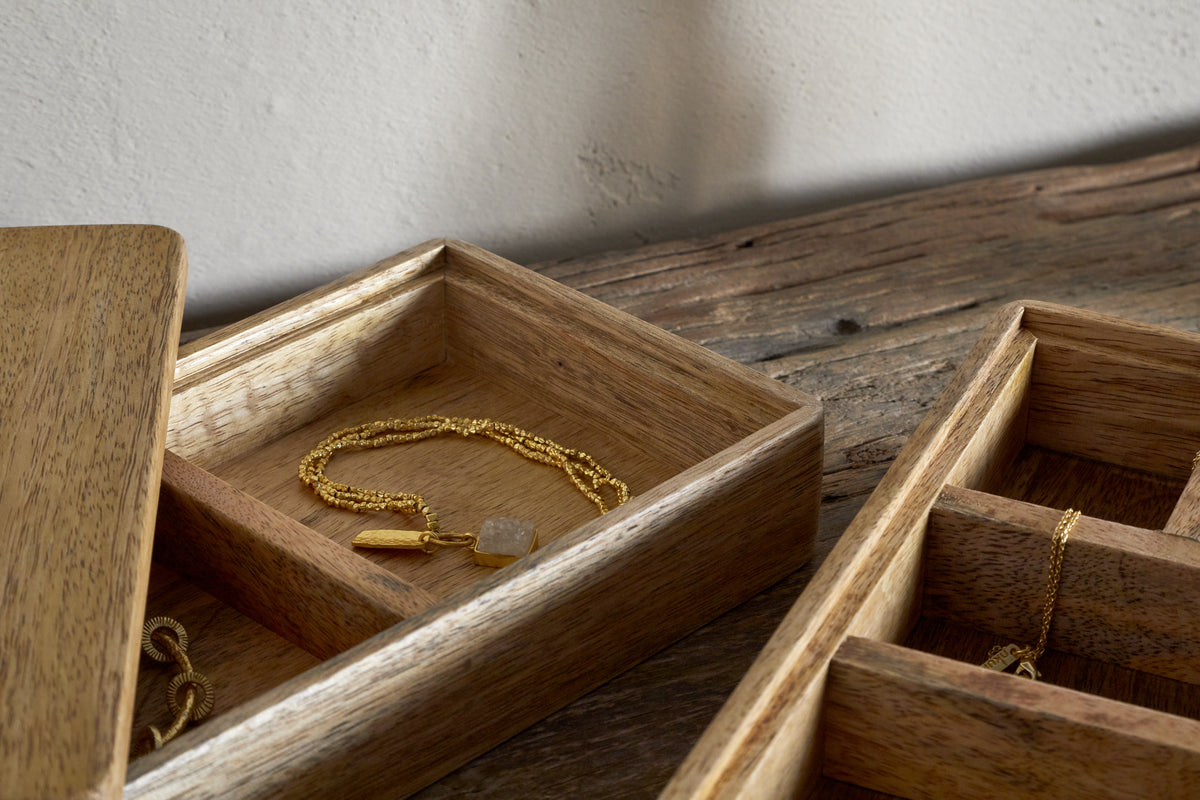 The nkuku Chanya Mango Wood Jewellery Box - Natural, with compartments for treasures, sits open on a rustic surface displaying a gold chain pendant, its natural wood grain set against a softly textured wall.