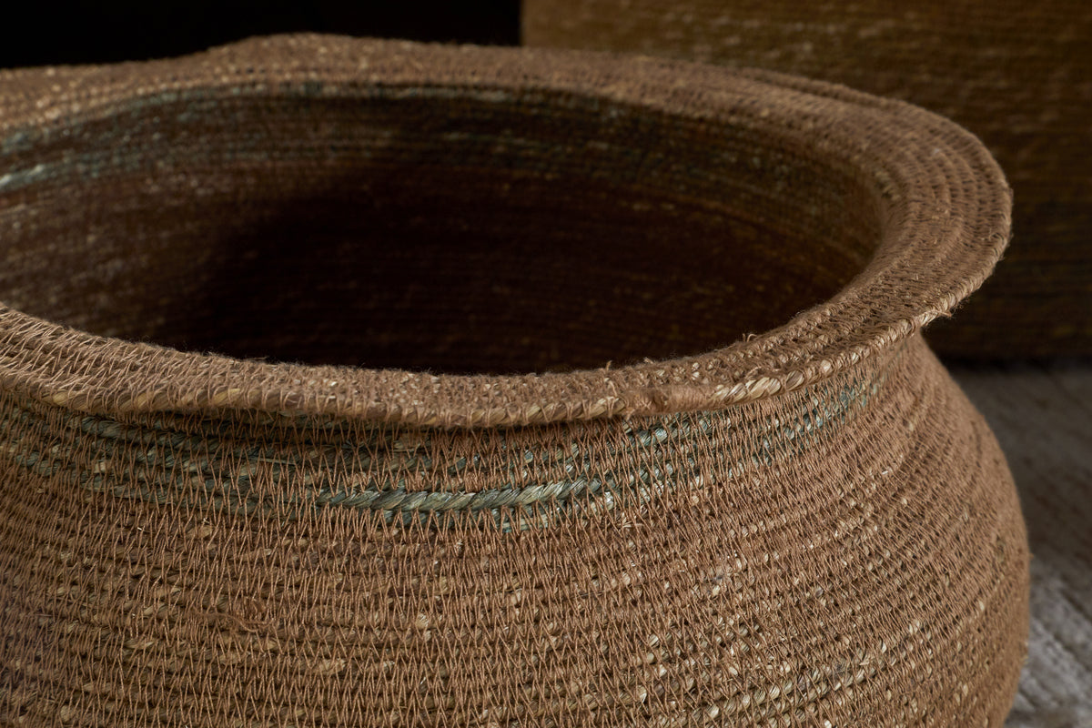 A close-up of the nkuku Rana Short Basket in Natural & Moss shows its handwoven texture, wide mouth, and thick rim. Coarse fibers with subtle green tones enhance its natural appeal. A similar basket appears partly in the background on light wood.