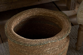 The nkuku Rana Tall Basket in Natural & Moss, featuring an uneven rim, sits empty on a textured light rug near wooden furniture and an upholstered chair.
