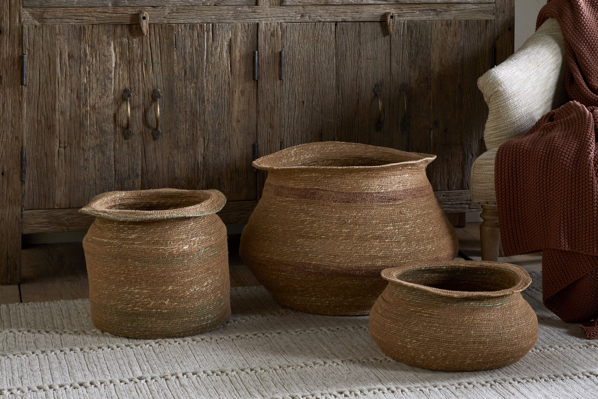 Three woven natural-fiber baskets, including the nkuku Rana Tall Basket in Natural & Moss, rest on a textured beige rug before a rustic wooden cabinet. On the right, a cream-upholstered chair and brown knit blanket are partly visible.