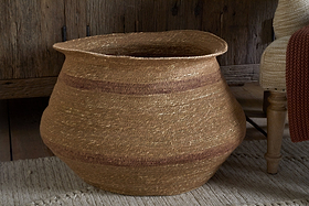 The nkuku Rana Wide Basket in Natural & Rust, featuring handwoven seagrass with subtle brown stripes, sits on a beige textured rug beside a wooden cabinet and upholstered chair, adding rustic warmth to the space.