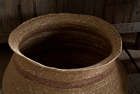 The nkuku Rana Wide Basket in Natural & Rust is a large, round seagrass storage basket featuring subtle horizontal stripes and earthy brown tones, shown on a wooden floor with a rustic cabinet in the background.