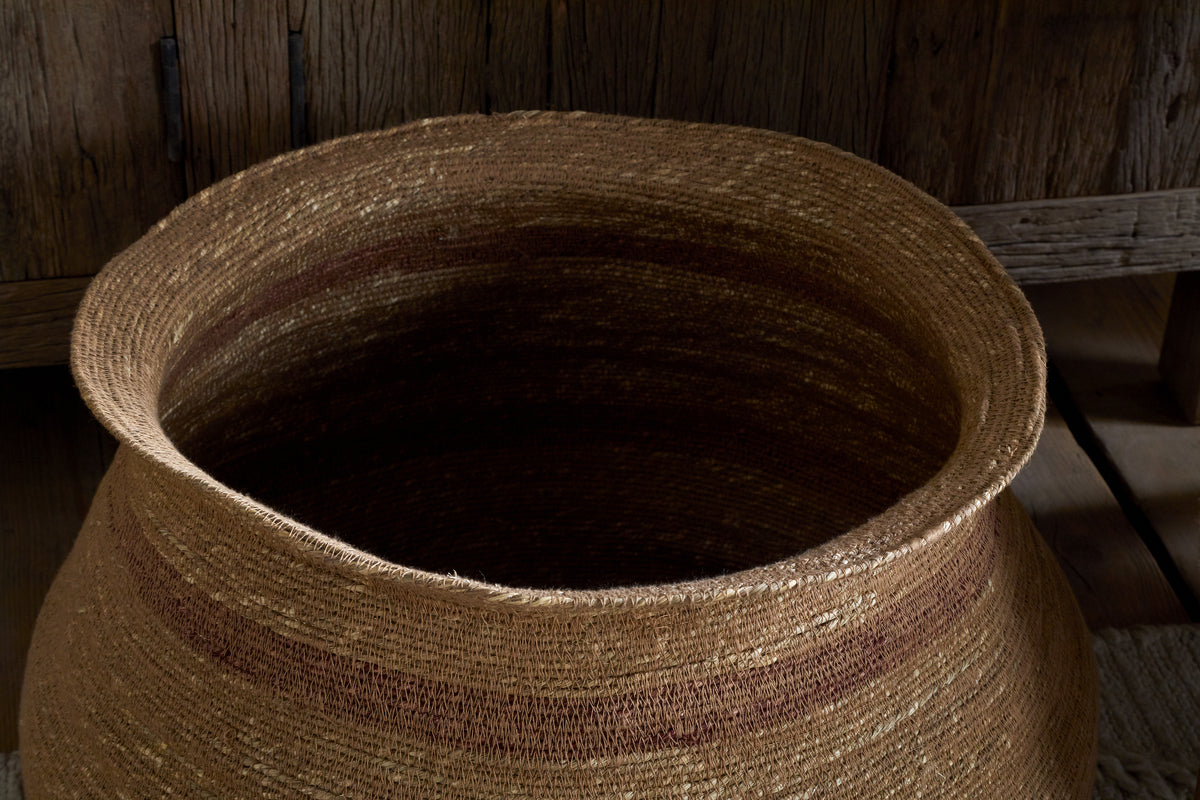 The nkuku Rana Wide Basket in Natural & Rust is a large, round seagrass storage basket featuring subtle horizontal stripes and earthy brown tones, shown on a wooden floor with a rustic cabinet in the background.