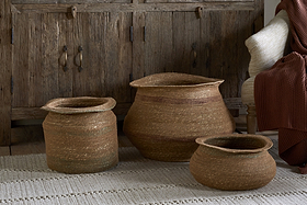 Three Rana Wide Baskets by nkuku in natural and rust tones are arranged on a cream rug before a rustic cabinet. A cream chair with a rust knit blanket adds warmth to this inviting storage basket display.
