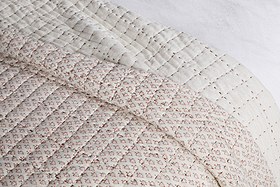 A close-up of nkuku's Peplu Cotton & Linen Screen Print Bed Quilt - Rust draped over a bed highlights its soft, textured finish with a rust-toned pattern on top and an off-white underside featuring subtle stitched lines.