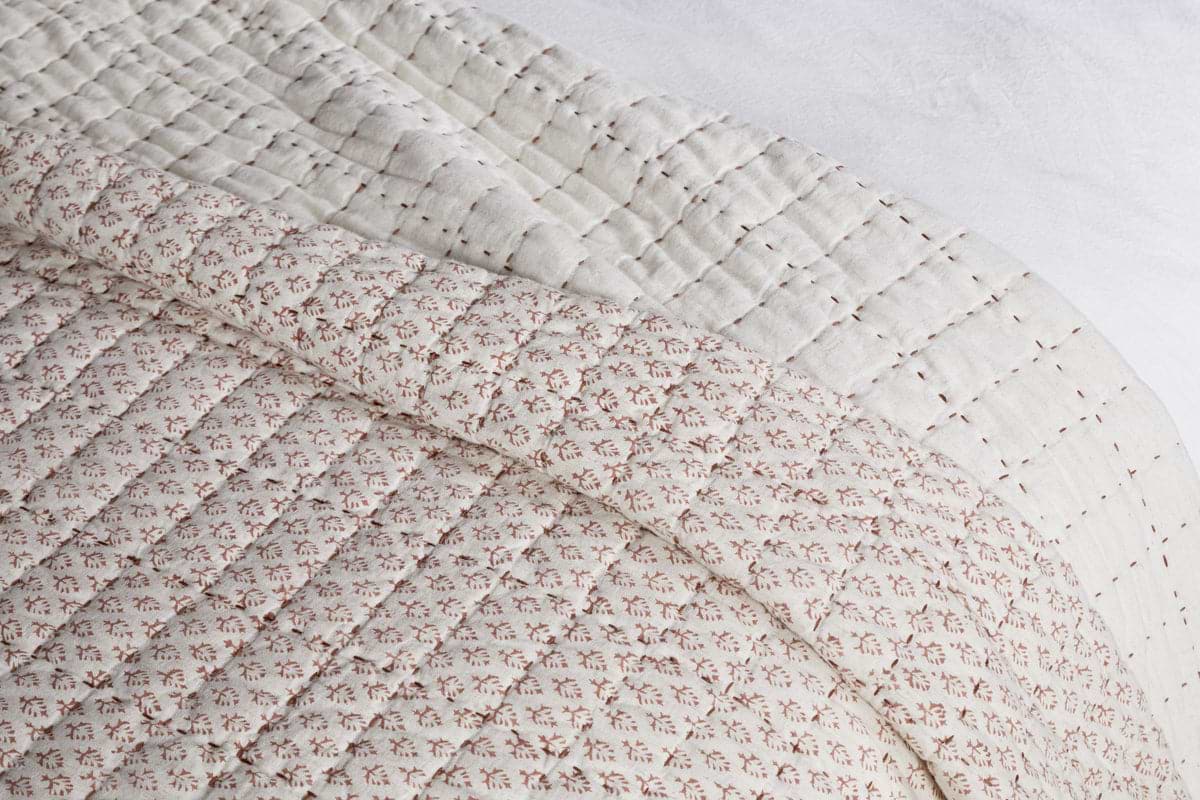 A close-up of nkuku's Peplu Cotton & Linen Screen Print Bed Quilt - Rust draped over a bed highlights its soft, textured finish with a rust-toned pattern on top and an off-white underside featuring subtle stitched lines.
