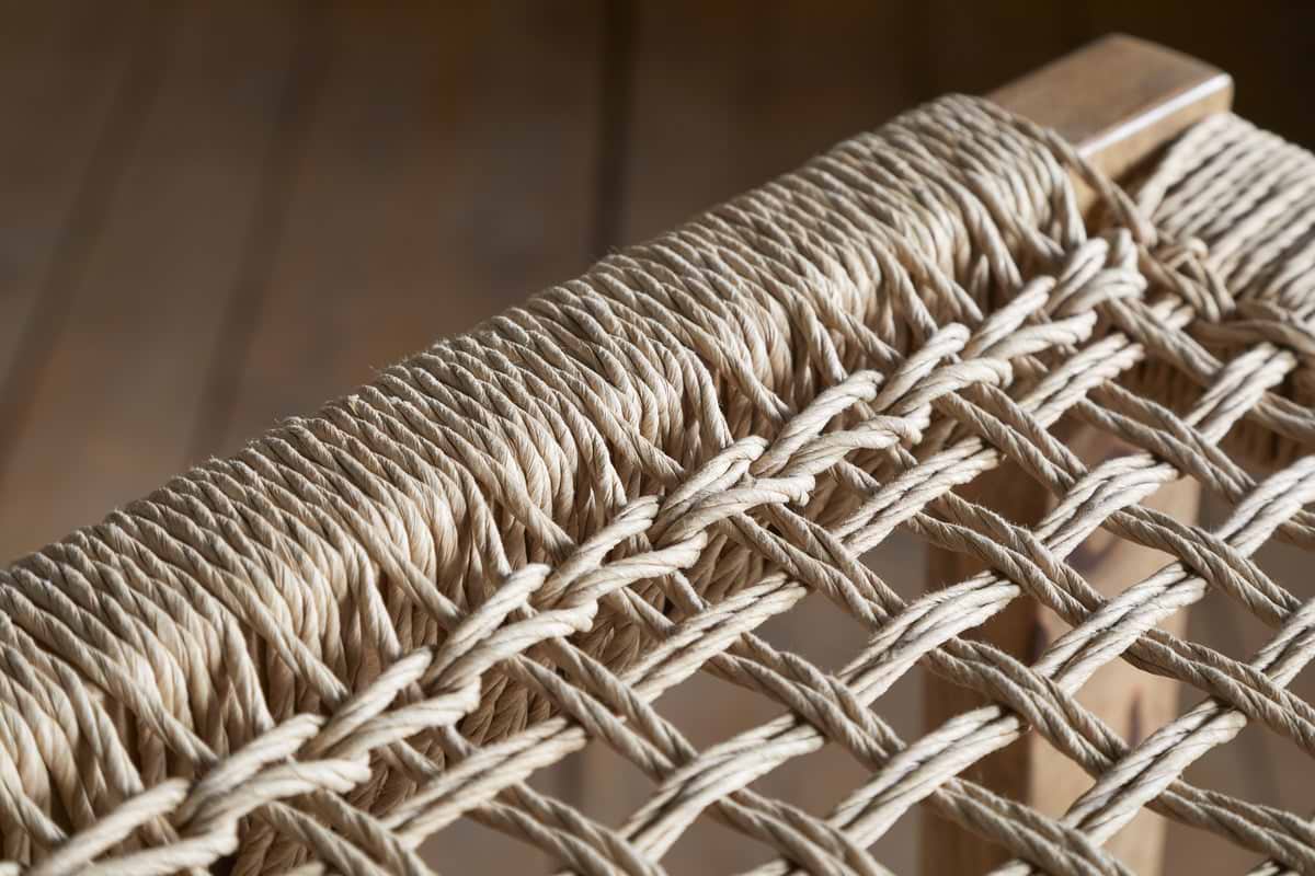 Close-up of the nkuku Aamani Mango Wood & Cord Counter Stool - Natural, featuring a tightly woven natural-colored rope seat on a smooth mango wood frame. Blurred wooden flooring appears in the background.