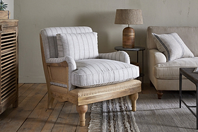 A cozy living room features the nkuku Abe Deconstructed Linen Armchair in natural stripe, boasting a sturdy mango wood frame and light gray cushions. A small round table with a brown lamp, beige sofa, and fringed textured rug complete the look.