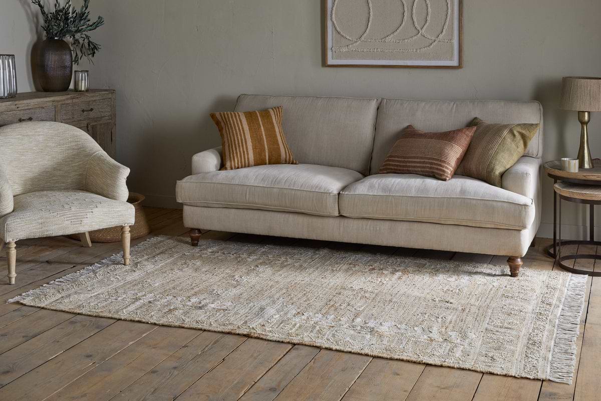 A cozy living room features a beige sofa with brown and green cushions, a nkuku Aqil Jute & Cotton Rug in natural tones on wooden floors, a light armchair, wooden side table, rustic cabinet with vases and greenery, and abstract art on neutral walls.