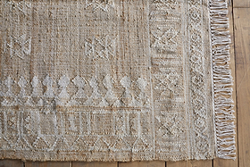 A close-up of the nkuku Aqil Jute & Cotton Rug - Natural reveals woven beige and white geometric patterns, a decorative border with fringed tassels, and highlights its rustic Indian design atop a wooden floor.