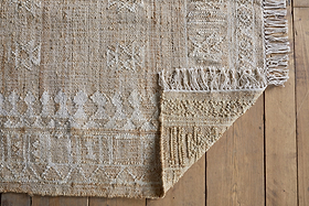 The nkuku Aqil Jute & Cotton Rug - Natural, in beige with white geometric patterns and knotted fringe, lies on a wooden floor. One corner is folded over, showing its thick weave and traditional Indian craftsmanship.