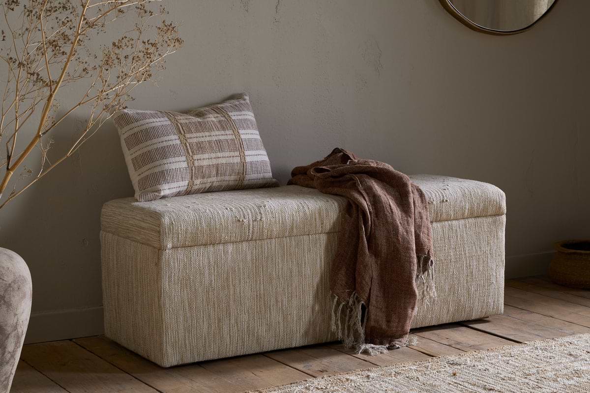 The Aranya Cotton & Jute Upholstered Storage Bench by nkuku sits against a light wall on a wooden floor, topped with a striped beige-and-white pillow and brown fringed throw. A round mirror, dried branches, and woven basket complete the cozy natural decor.