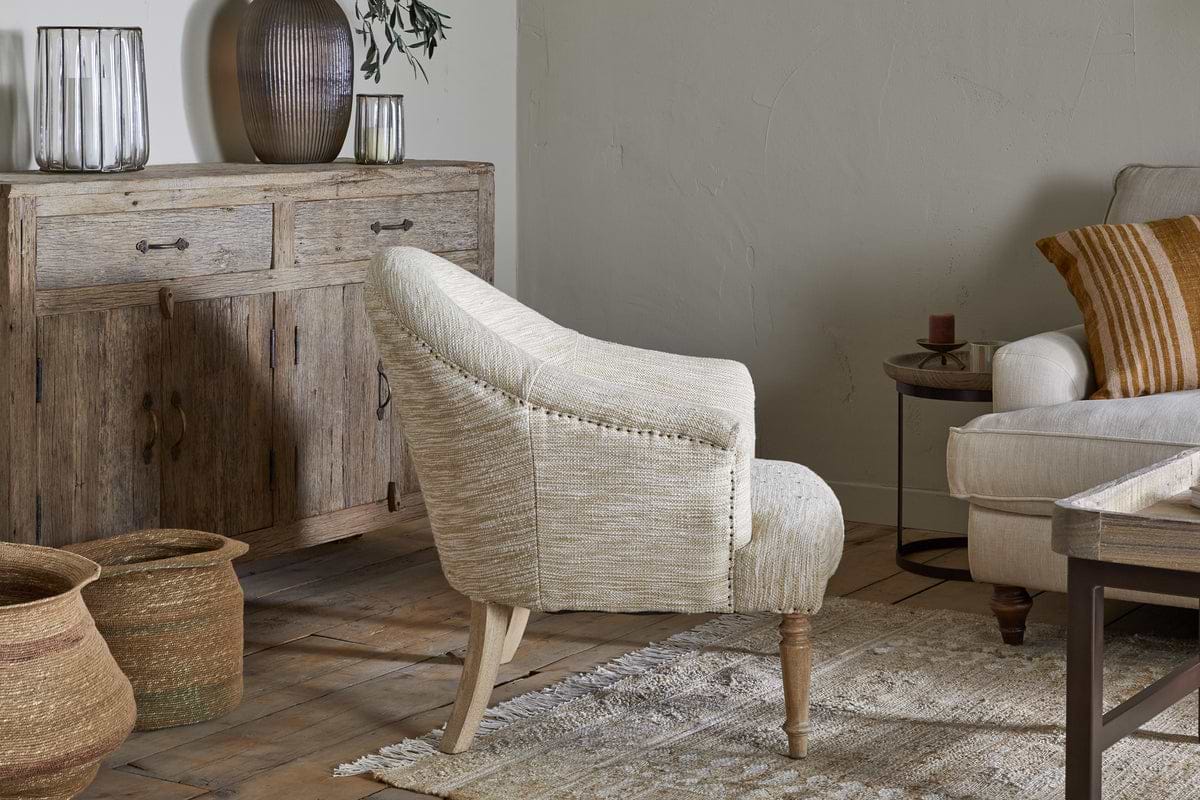 The nkuku Aranya Cotton & Jute Occasional Chair in natural light upholstery sits on a patterned rug in a rustic living room with wooden floors, a wooden sideboard, decorative vases, woven baskets, and a beige sofa with striped pillows.