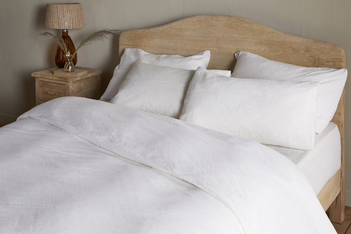 A neatly made bed features the Cela Hemp & Cotton Duvet Cover - Natural by nkuku, sustainable bedding, and four white pillows beside a wooden nightstand. A brown glass lamp with a beige shade and decorative twig enhances the light beige wall.