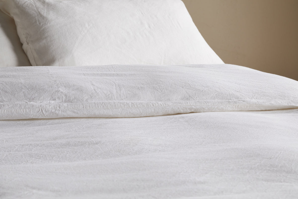 Close-up of a neatly made bed featuring the nkuku Cela Hemp & Cotton Duvet Cover in Natural, with white linen sheets and two pillows. The duvet's subtle texture and sustainable material create a soft, inviting look against the calming wall.