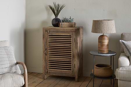 The Ibo Reclaimed Wood Slatted Low Cabinet by nkuku sits on a wooden floor with small plants in dark vases on top. A round side table holds a woven lamp and basket, framed by two beige armchairs for a cozy Indian craft-inspired scene.