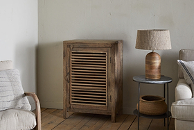 The Ibo Reclaimed Wood Slatted Low Cabinet by nkuku stands against a pale wall. Next to it, a round side table displays a woven lamp and basket. A beige sofa with a striped pillow and wooden chair arm rests on the wooden floor.
