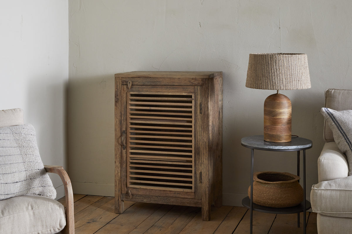 The Ibo Reclaimed Wood Slatted Low Cabinet by nkuku stands against a pale wall. Next to it, a round side table displays a woven lamp and basket. A beige sofa with a striped pillow and wooden chair arm rests on the wooden floor.