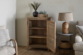 The nkuku Ibo Reclaimed Wood Slatted Low Cabinet - Natural stands in a cozy corner with open shelves. A black vase and plant tray sit on top. Nearby, a round table displays a woven lamp and basket beside a beige sofa and wood floor.