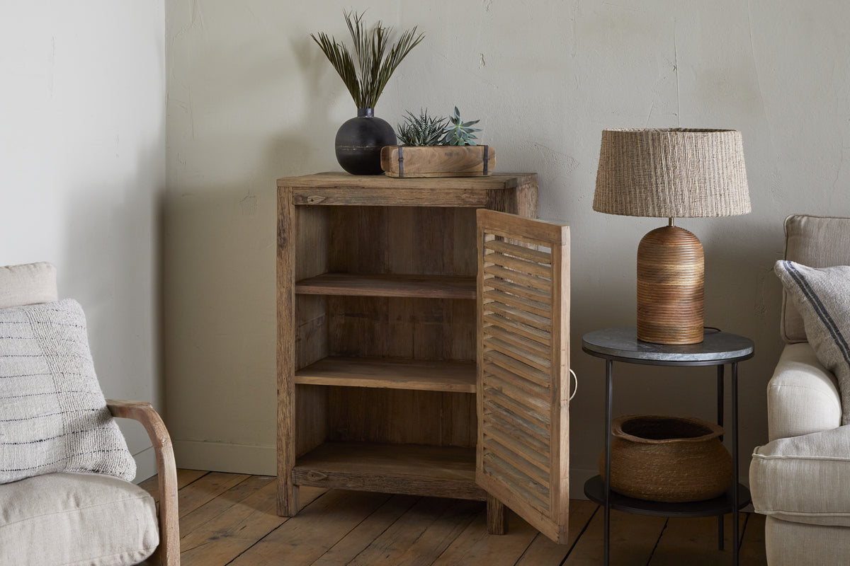 The nkuku Ibo Reclaimed Wood Slatted Low Cabinet - Natural stands in a cozy corner with open shelves. A black vase and plant tray sit on top. Nearby, a round table displays a woven lamp and basket beside a beige sofa and wood floor.
