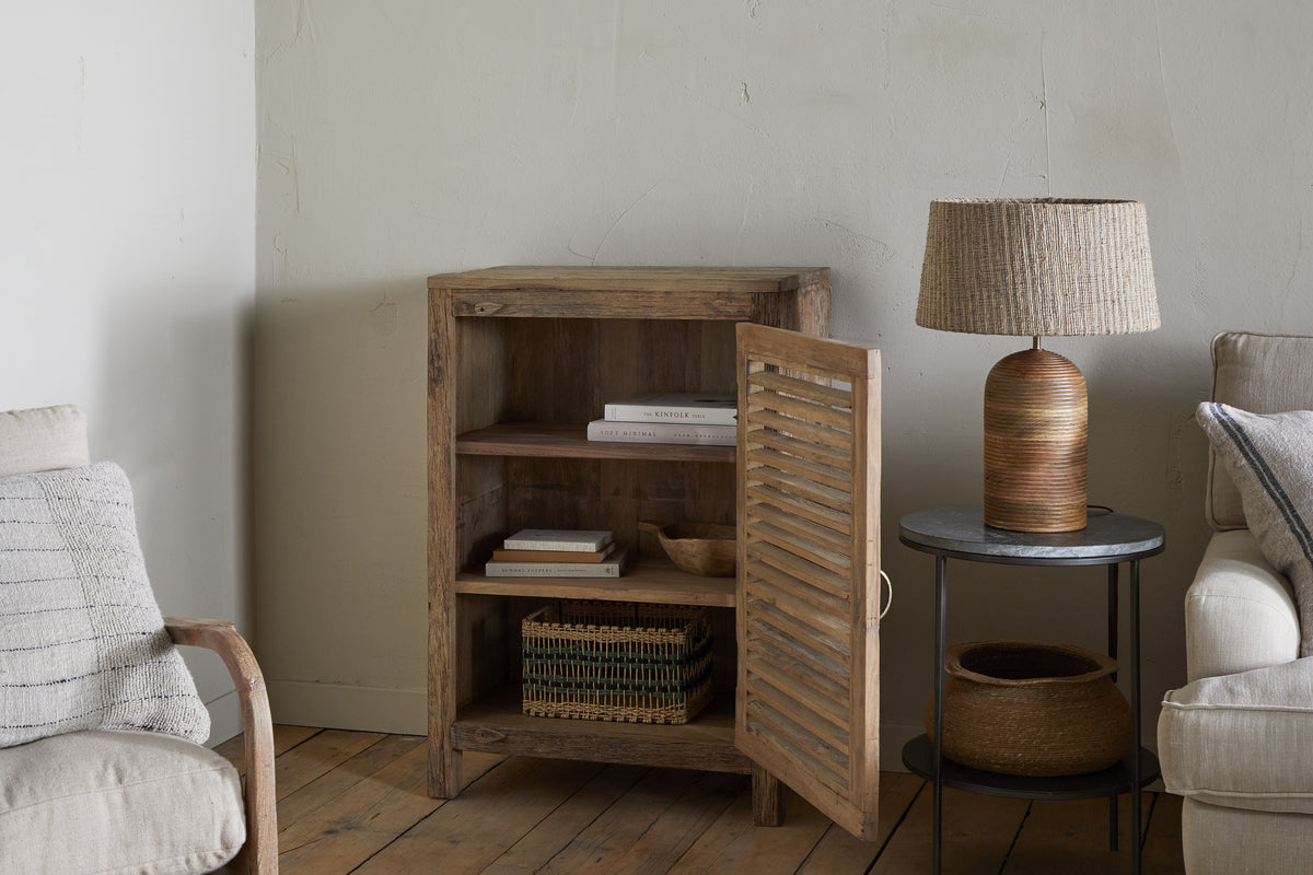 A cozy living room corner features the nkuku Ibo Reclaimed Wood Slatted Low Cabinet - Natural with its door open displaying books and baskets. Nearby, a round side table holds a woven lamp, and a beige armchair with wooden arms sits next to the cabinet.