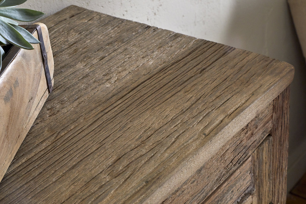 A close-up of the nkuku Ibo Reclaimed Wood Slatted Low Cabinet - Natural shows its textured, rustic surface and visible grain. A small wooden planter with a green plant appears in the corner, set against a slightly worn, neutral wall.
