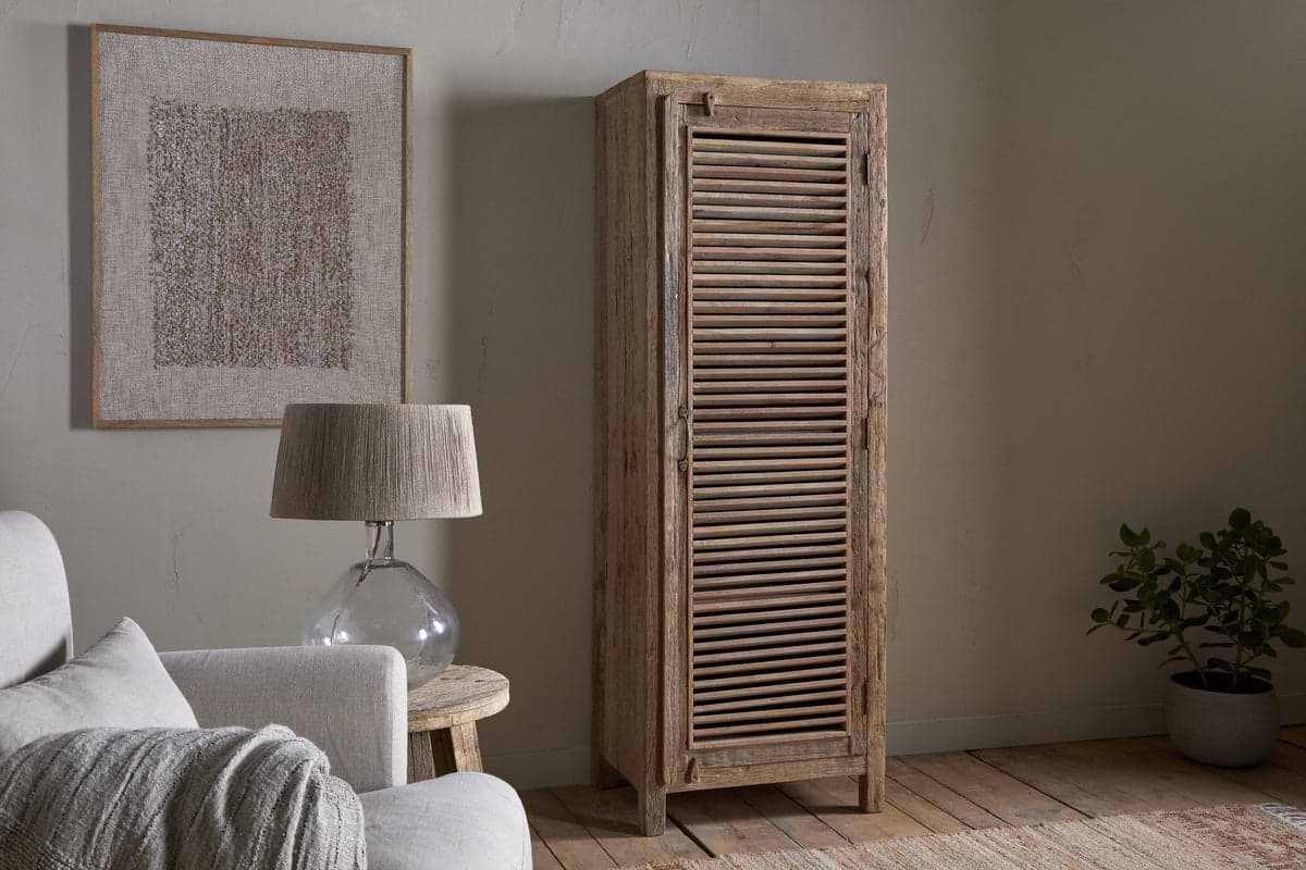 A cozy room features a light sofa, wooden side table with glass lamp, the nkuku Ibo Reclaimed Wood Slatted Tall Cabinet - Natural, a potted plant, neutral artwork on a beige wall, and warm wood floors for a natural minimalist look.