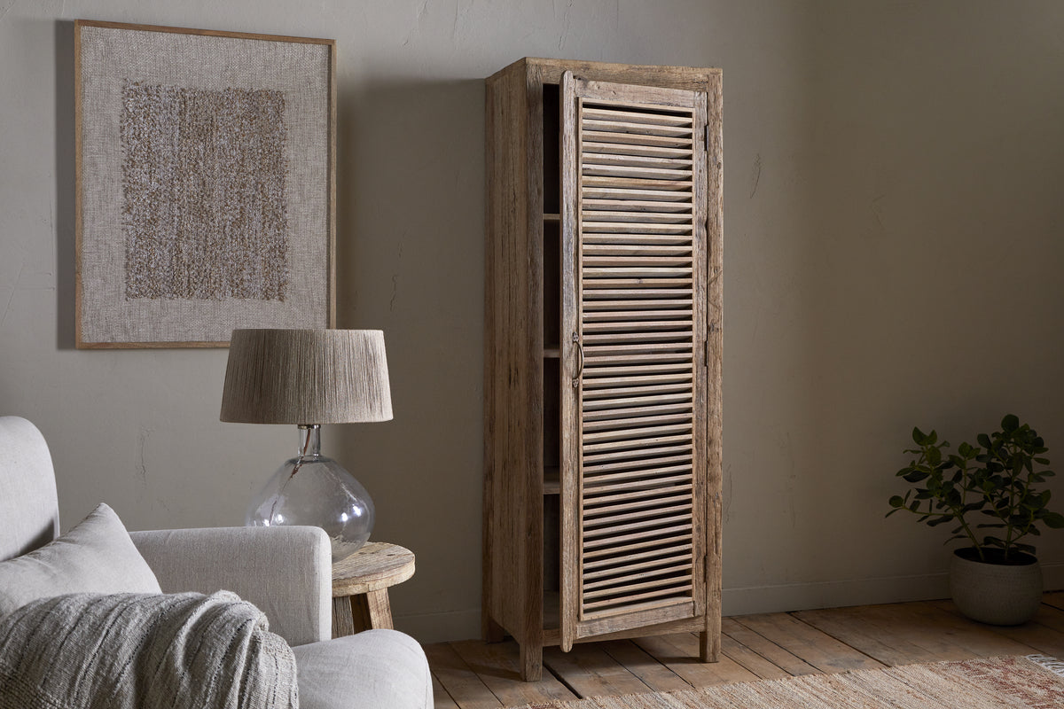 A cozy, neutral-toned living room features the nkuku Ibo Reclaimed Wood Slatted Tall Cabinet in natural, framed abstract artwork, a round glass lamp on a rustic side table, white sofa, light rug, potted plant, and wooden floor.