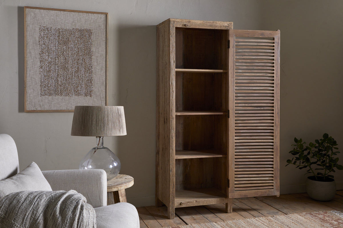The Ibo Reclaimed Wood Slatted Tall Cabinet by nkuku stands partially open in a cozy living room with a light sofa, glass-based table lamp, framed textured artwork, a potted plant, and wooden floors.
