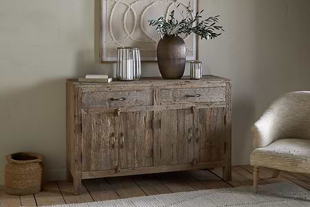 The nkuku Ibo Reclaimed Wood Sideboard – Natural, with two drawers and cabinet doors, sits against a beige wall. A brown vase with leafy branches, glass candle holders, books, and cozy accents add to the neutral-toned room.