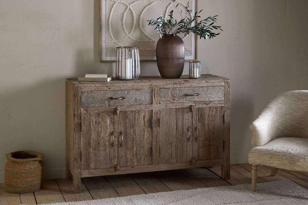The nkuku Ibo Reclaimed Wood Sideboard – Natural, with two drawers and cabinet doors, sits against a beige wall. A brown vase with leafy branches, glass candle holders, books, and cozy accents add to the neutral-toned room.