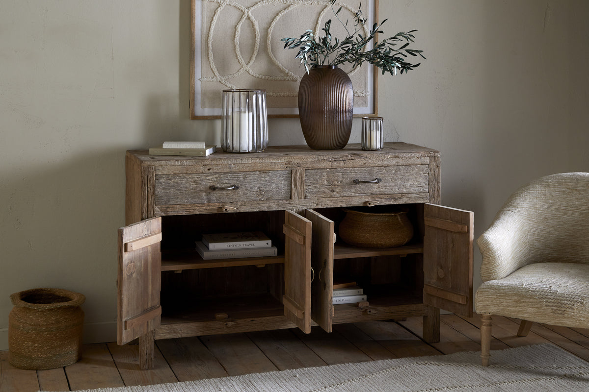The nkuku Ibo Reclaimed Wood Sideboard - Natural exudes rustic charm with two drawers and four open doors showing shelves. On top are books, a brown vase with green branches, and a glass lantern. A woven basket sits by a cream upholstered chair.