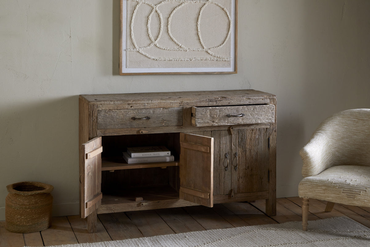 A rustic vibe fills the room with the nkuku Ibo Reclaimed Wood Sideboard - Natural, one drawer open and both cabinet doors ajar to reveal books. It stands on a wooden floor beside a woven basket and curved cream chair beneath abstract artwork.