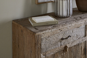 The Ibo Reclaimed Wood Sideboard - Natural by nkuku features a weathered texture, simple metal handle, and earthy neutral tones. It holds two stacked books, a lamp with a ribbed glass base, and part of a brown vase for rustic charm.