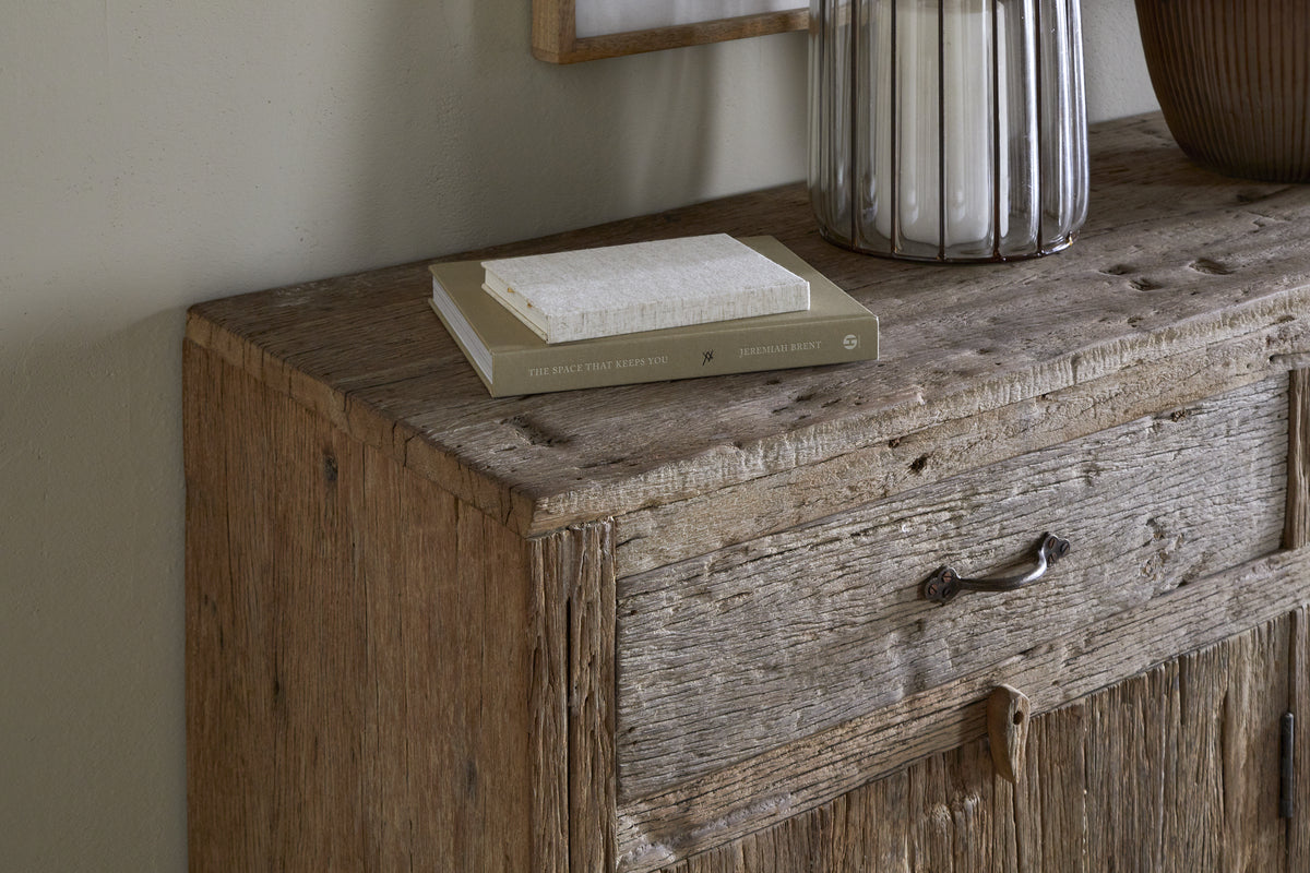 The Ibo Reclaimed Wood Sideboard - Natural by nkuku features a weathered texture, simple metal handle, and earthy neutral tones. It holds two stacked books, a lamp with a ribbed glass base, and part of a brown vase for rustic charm.