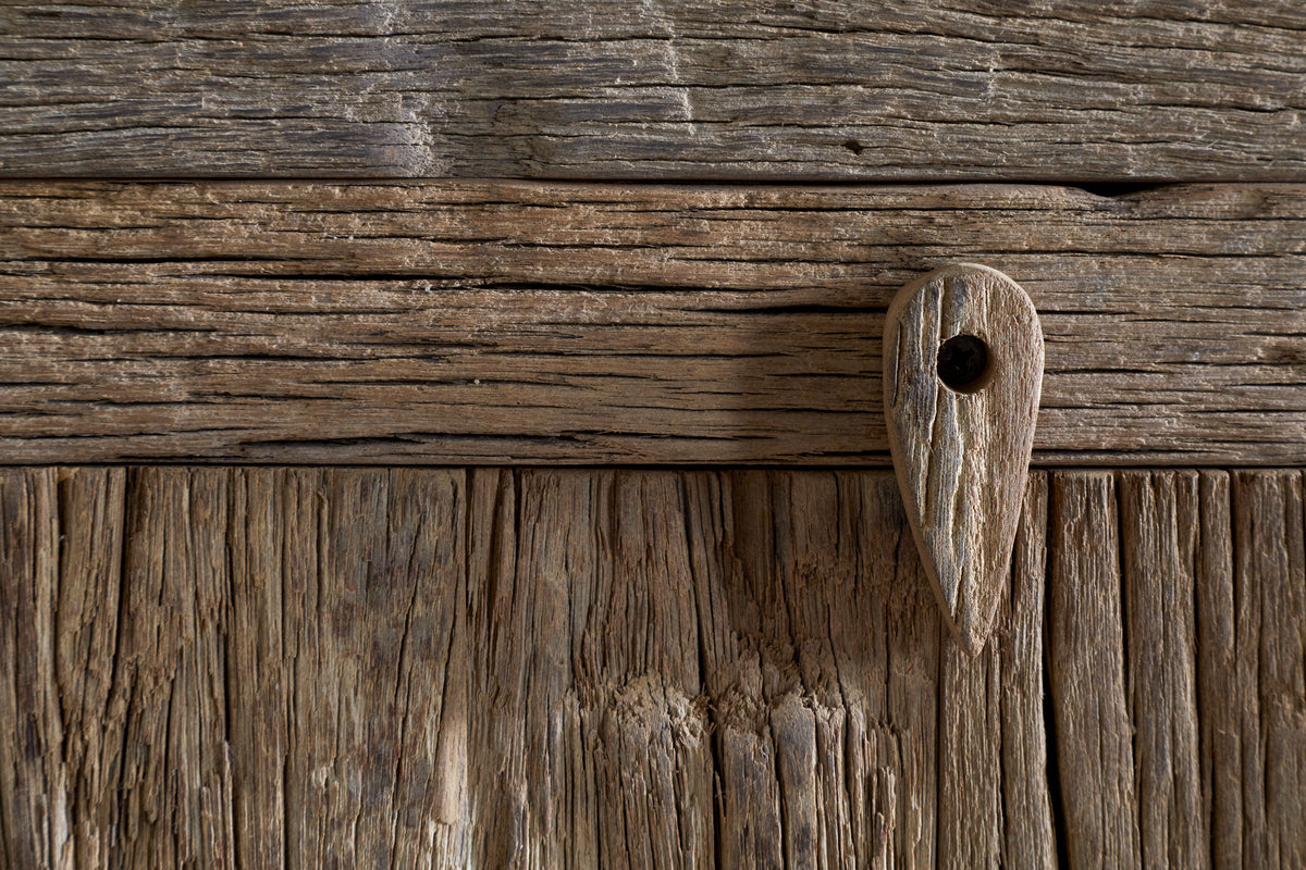 Close-up of a weathered wooden door with textured planks, featuring a tear-shaped wooden peg and dark screw—showcasing the rustic charm and natural patterns found in the nkuku Ibo Reclaimed Wood Sideboard - Natural.
