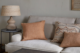 A beige sofa is adorned with two rust-striped cushions and the Ilaa Wool Cushion Cover - Sand by nkuku, featuring a geometric texture. A round wooden side table with a woven lamp stands beside it, against a subtly cracked light gray wall for rustic charm.