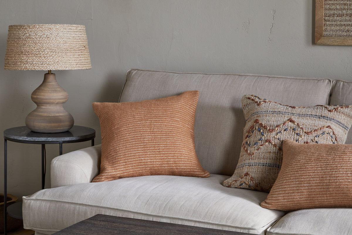 A beige sofa is adorned with two rust-striped cushions and the Ilaa Wool Cushion Cover - Sand by nkuku, featuring a geometric texture. A round wooden side table with a woven lamp stands beside it, against a subtly cracked light gray wall for rustic charm.