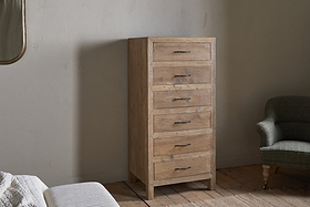 The nkuku Iya Reclaimed Wood Tall Boy Chest of Drawers - Natural, with six drawers and black metal handles, stands against a light wall, complemented by a green upholstered armchair, white bed, and wooden floor for a cozy, rustic feel.