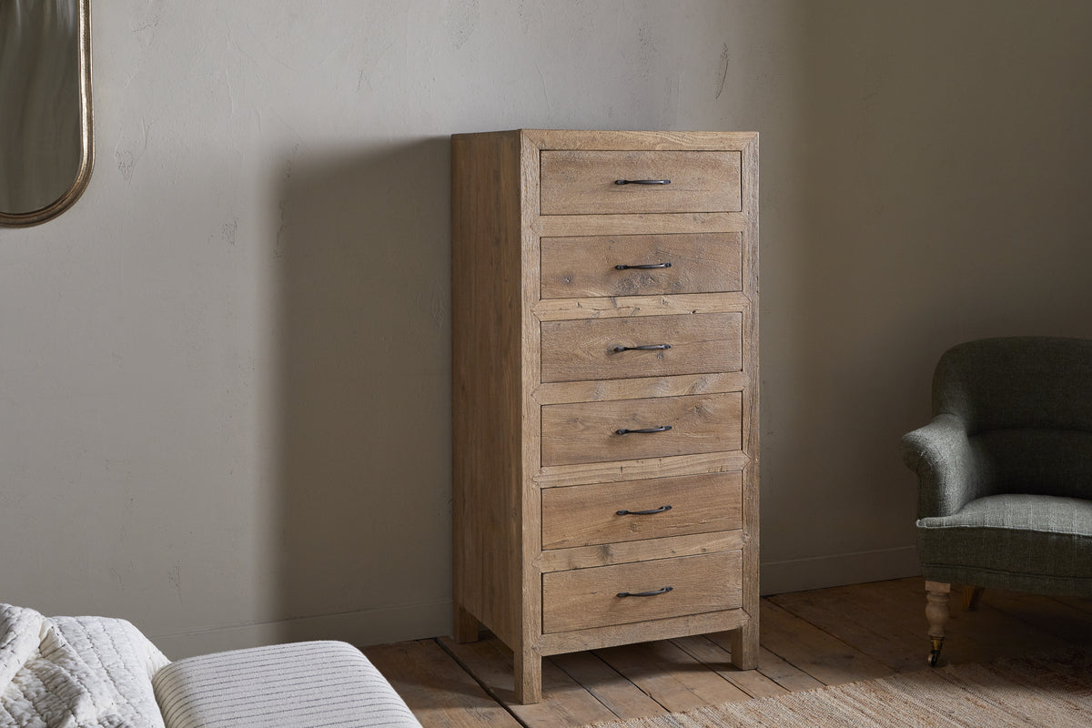 The nkuku Iya Reclaimed Wood Tall Boy Chest of Drawers - Natural, with six drawers and black metal handles, stands against a light wall, complemented by a green upholstered armchair, white bed, and wooden floor for a cozy, rustic feel.