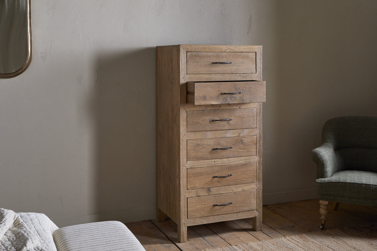 The Iya Reclaimed Wood Tall Boy Chest of Drawers by nkuku, featuring six drawers (one slightly open), stands on wooden floors beside a green armchair and a partially visible bed, set against textured light walls with soft natural lighting.