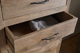 A close-up of the nkuku Iya Reclaimed Wood Tall Boy Chest of Drawers - Natural shows a partially open drawer with a neatly folded gray towel inside, textured finish, black metal handles, set on a wooden floor against neutral-toned walls.