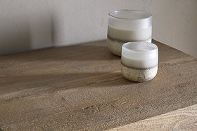 Two frosted glass candles with white wax and rustic metallic bases sit on the Iya Reclaimed Wood Tall Boy Chest of Drawers - Natural by nkuku, set against a neutral wall for a minimal look.
