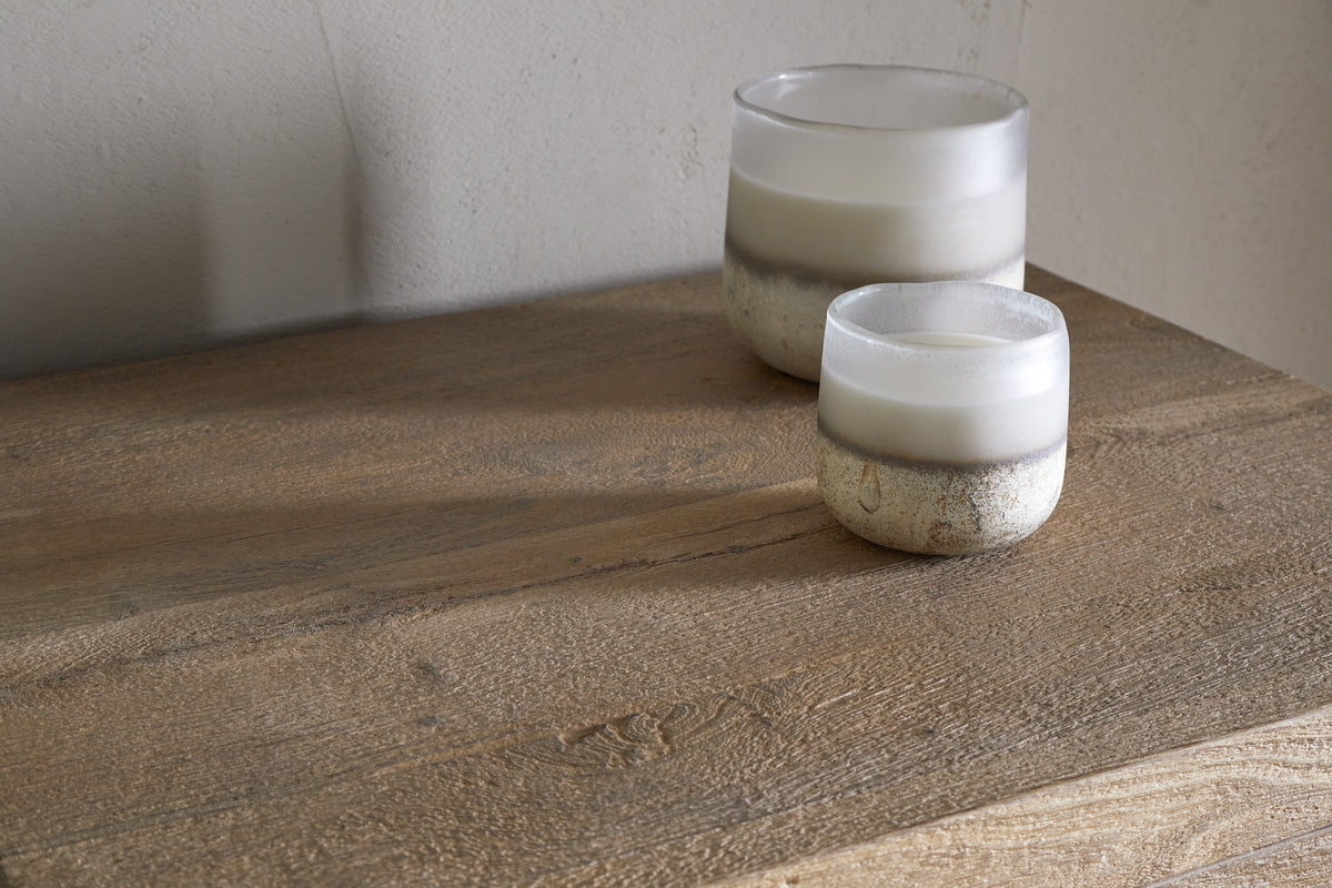 Two frosted glass candles with white wax and rustic metallic bases sit on the Iya Reclaimed Wood Tall Boy Chest of Drawers - Natural by nkuku, set against a neutral wall for a minimal look.