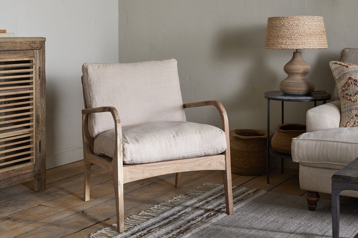 A cozy living room with the nkuku Jaya Acacia Wood & Linen Occasional Chair in natural, a rustic wooden cabinet, side table with woven lamp, two baskets, gray sofa with patterned pillow, and striped rug on wooden floorboards.
