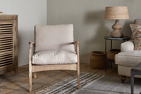 A cozy living room with nkuku’s Jaya Acacia Wood & Linen Occasional Chair - Natural, a striped rug on wooden floors, a wooden cabinet, beige sofa with linen cushions, two woven baskets, round side table, and lamp with woven shade against a light wall.